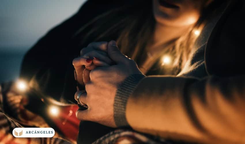 Manos entrelazadas de una pareja iluminadas por luces cálidas, representando conexión, amor y devoción espiritual en el contexto de la novena al Arcángel para el amor