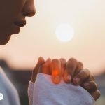 Persona orando con las manos juntas al amanecer, simbolizando fe y conexión espiritual en momentos de búsqueda de salud y protección divina.