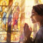 Mujer rezando en una iglesia con luz suave entrando por vitrales, reflejando figuras de arcángeles.
